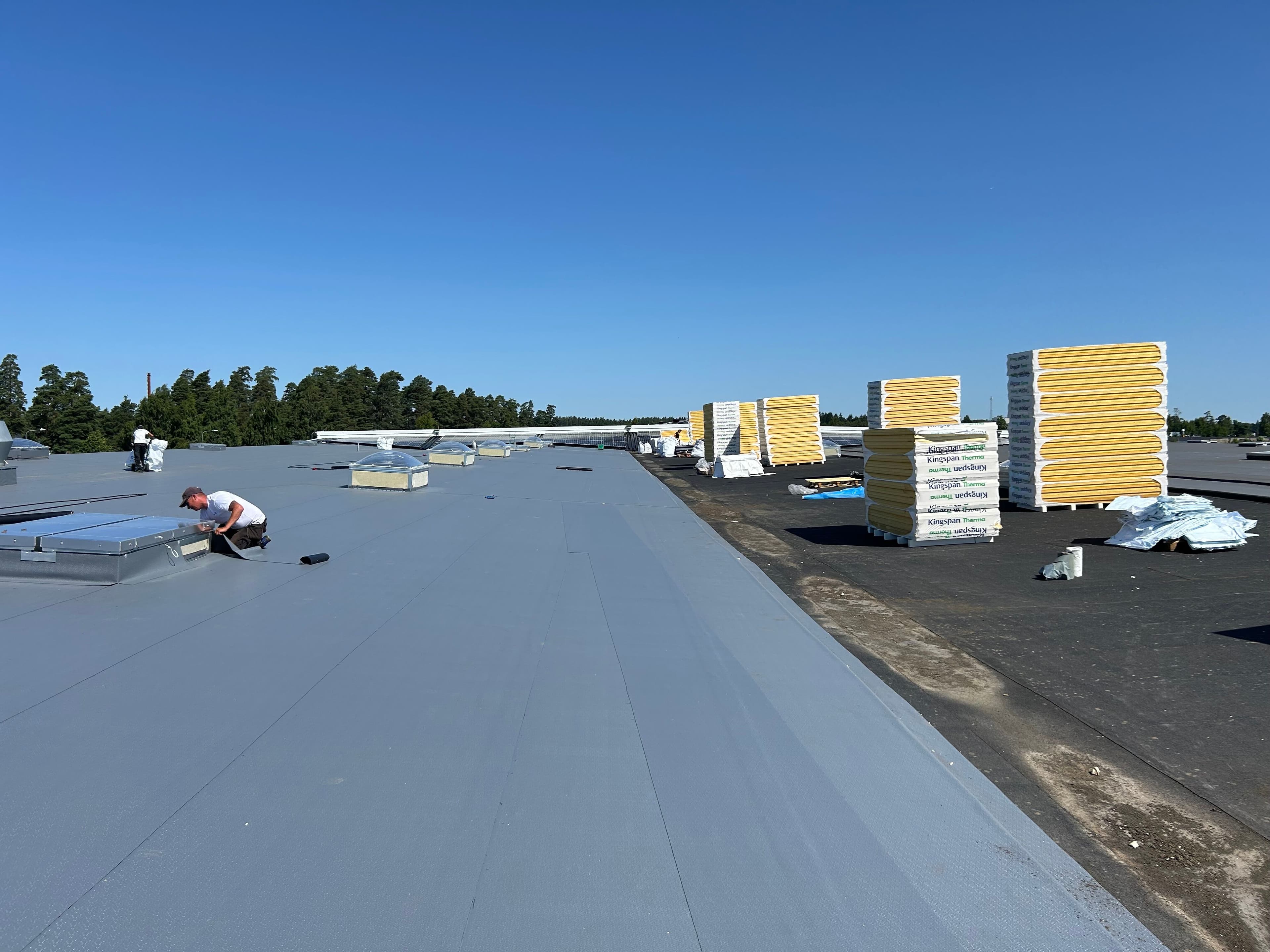 Takläggare arbetar med takrenovering på ett platt tak där Kingspan Therma isolerskivor används för tilläggsisolering. Staplar med isolerskivor syns på taket under pågående installation.