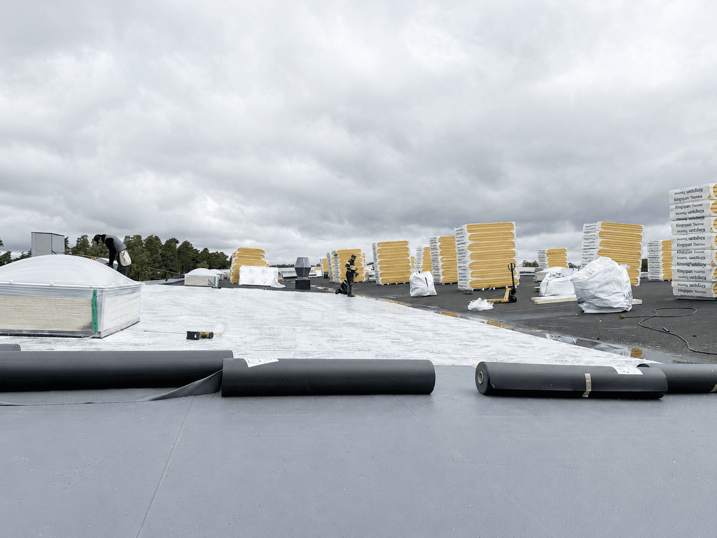 Kingspan Therma isolerskivor installeras på ett platt tak under en takrenovering. Takläggare arbetar med isoleringen medan rullar med takduk, takfönster och staplade isolerskivor syns på taket.