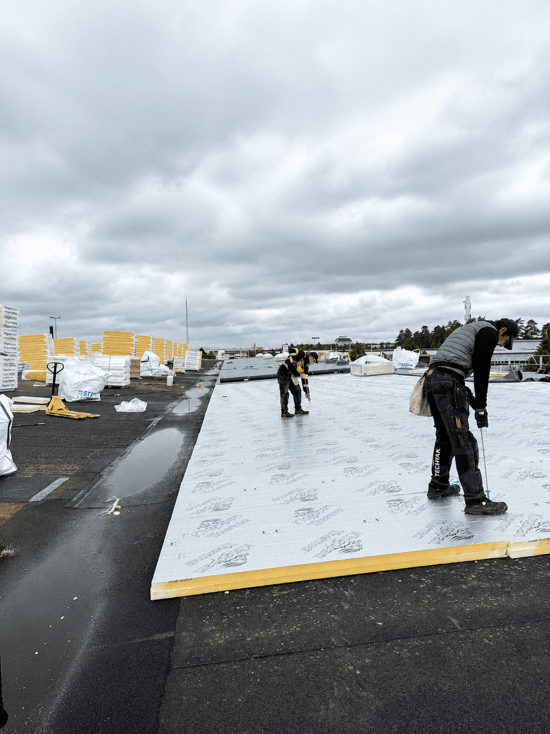 Takläggare installerar Kingspan Therma isolerskivor på ett platt tak under en takrenovering. Isolerskivorna mäts och anpassas inför installation medan fler skivor och återvinningssäckar syns i bakgrunden.
