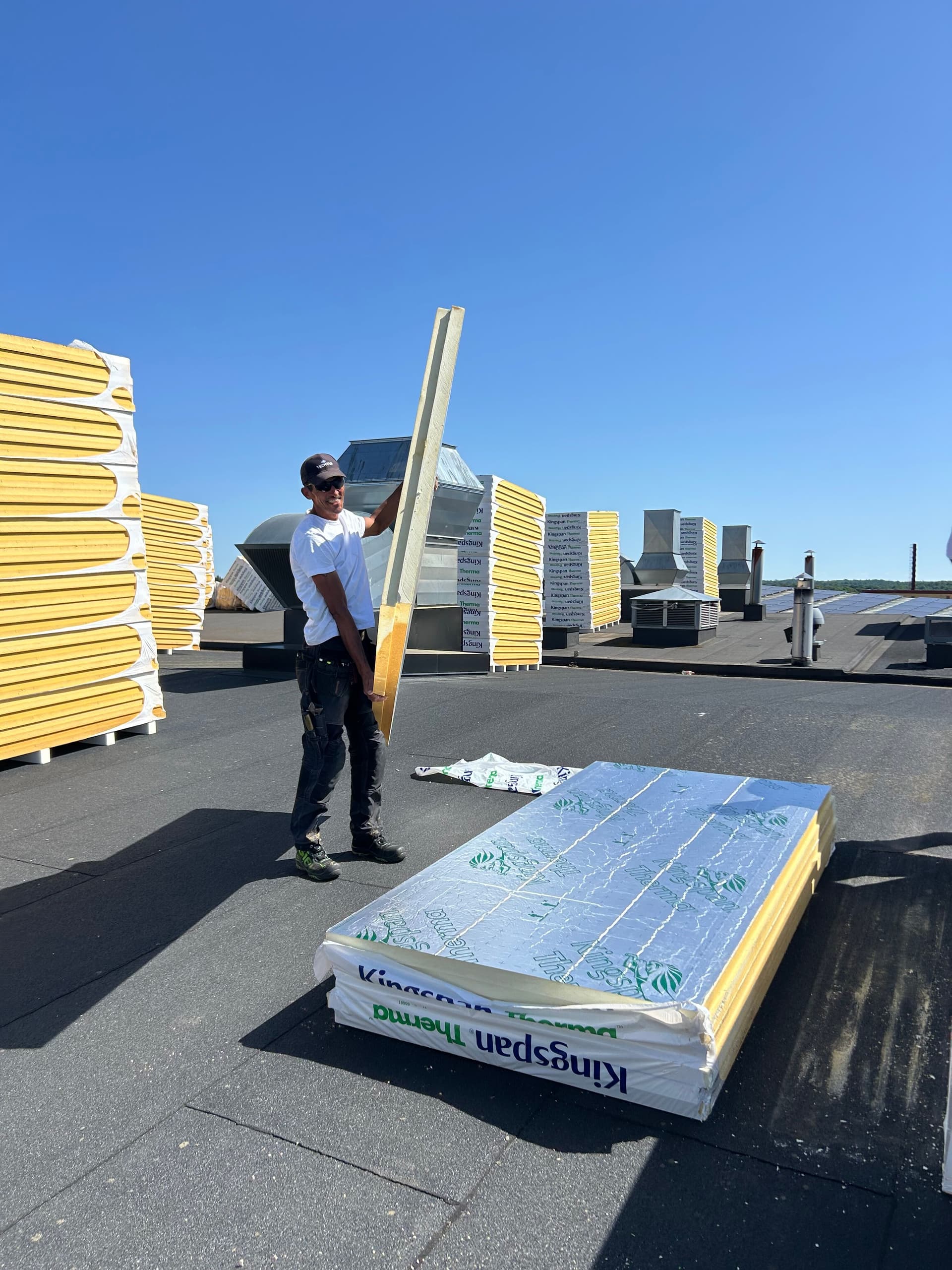 Takläggare bär en Kingspan Therma isolerskiva på ett platt tak under en takrenovering. Flera staplar med isolerskivor syns på taket inför den fortsatta installationen.